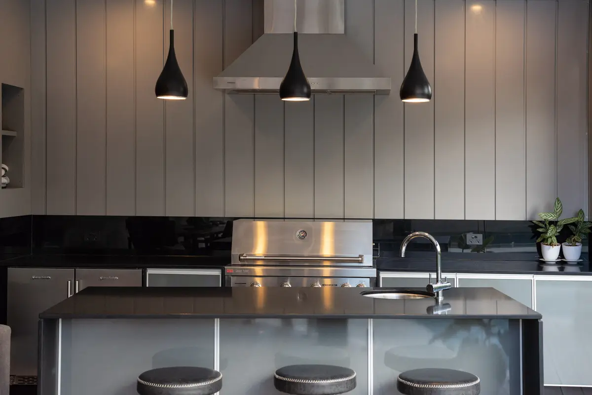 Barbecue and stainless range hood set into a grey clad wall, pendant lights over the island bench