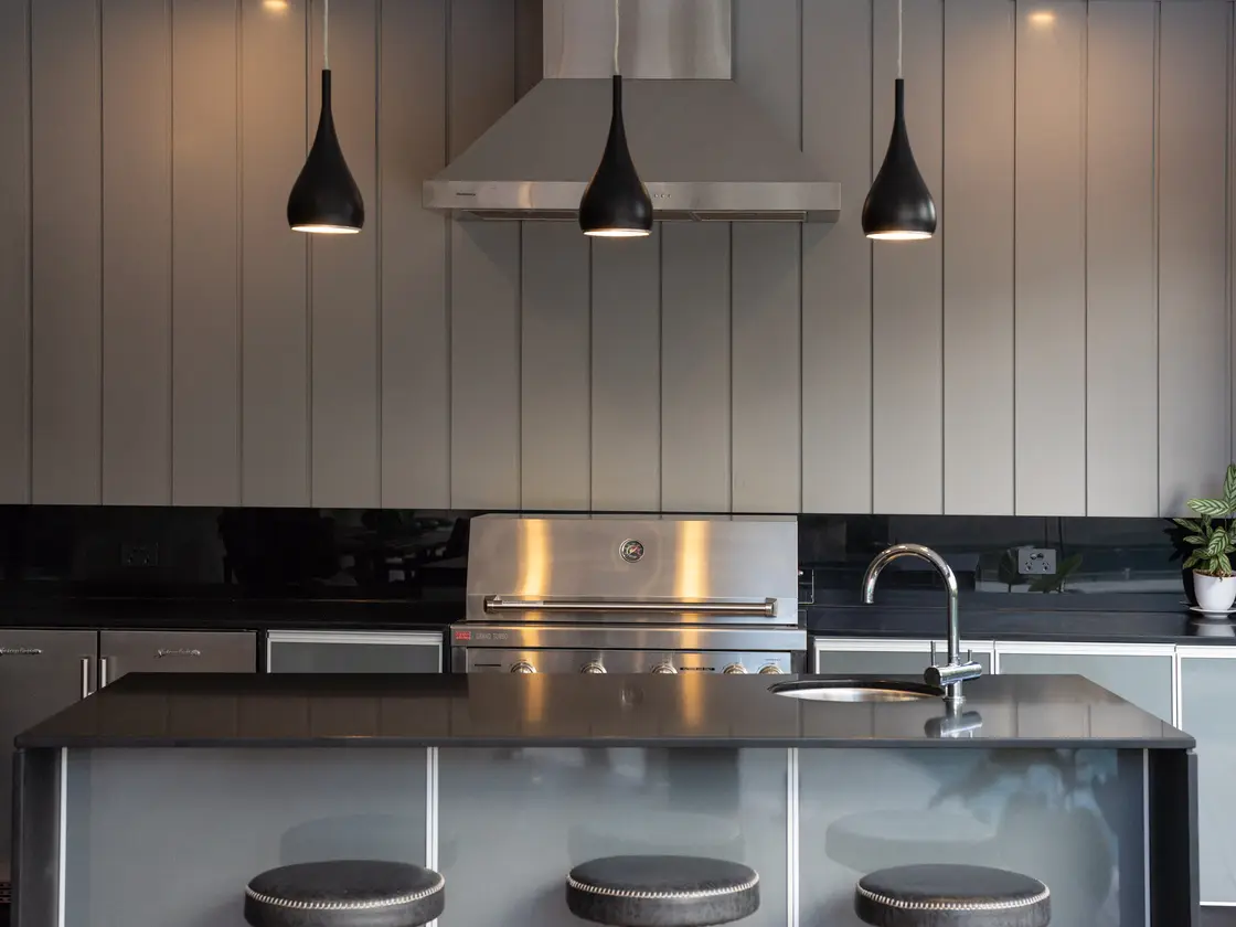 Full-height pale grey clad wall behind a built-in barbecue and stainless range hood, pendant lights over the island bench