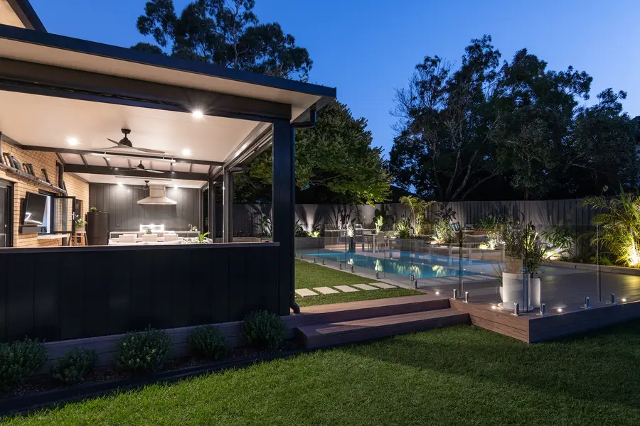 The lit alfresco and outdoor kitchen at dusk, seen across the lawn past the pool