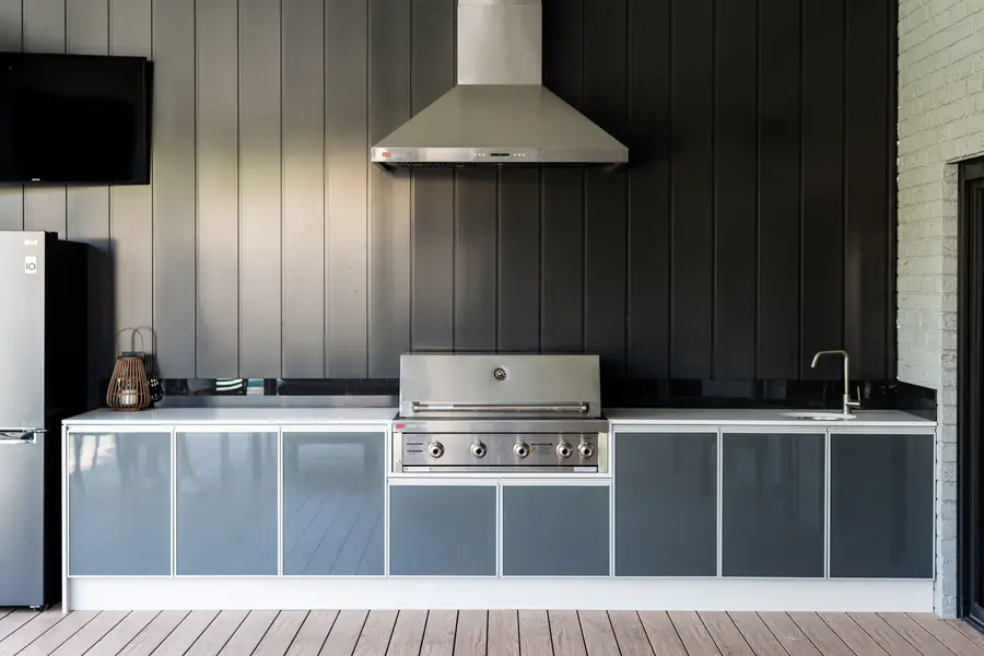 Outdoor kitchen with a stainless range hood set into the dark clad wall, beside a wall-mounted TV