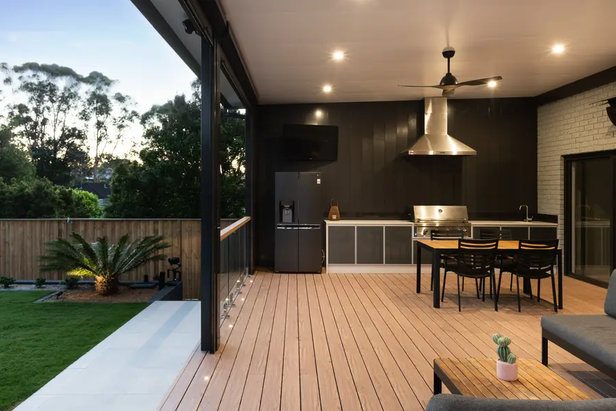 The outdoor kitchen and dining end of the deck at dusk, with the clad wall lit behind