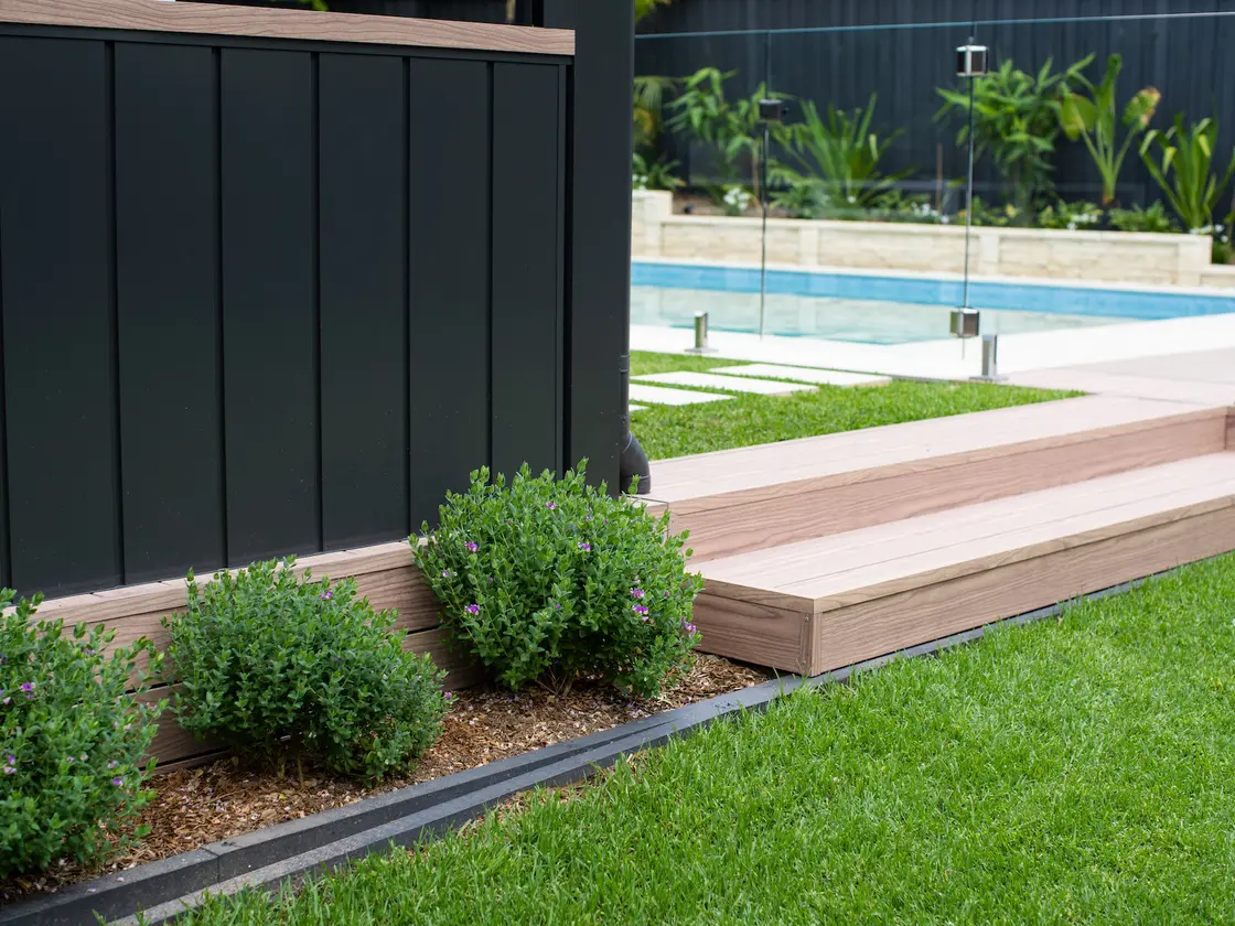 Low dark powder-coated clad wall with timber-look capping, running along the edge of the deck beside the pool