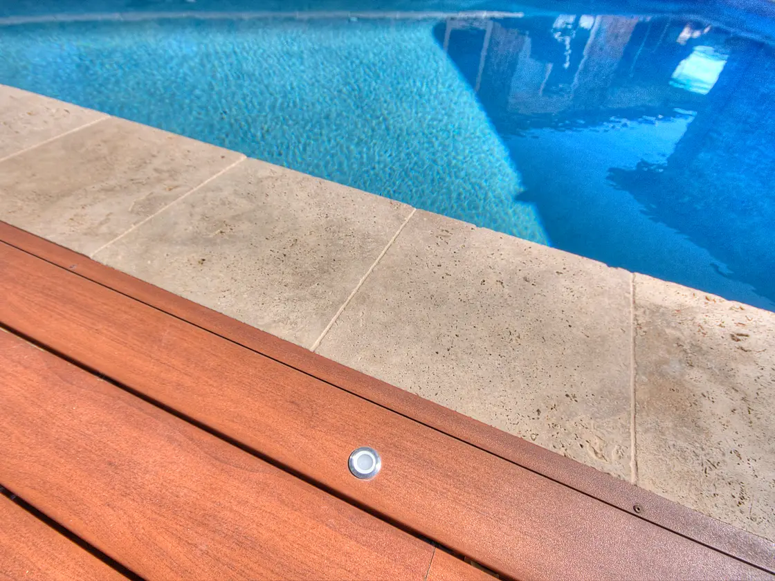 Small round in-floor LED set flush into a reddish-brown deck board beside the pool coping, lit water behind