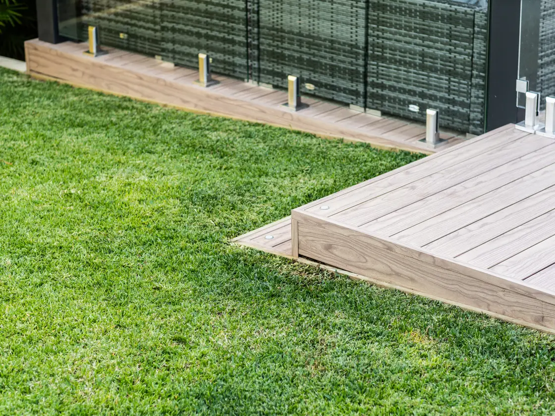 Pale timber-look aluminium deck boards meeting the lawn at a clean mitred edge, glass pool fence behind