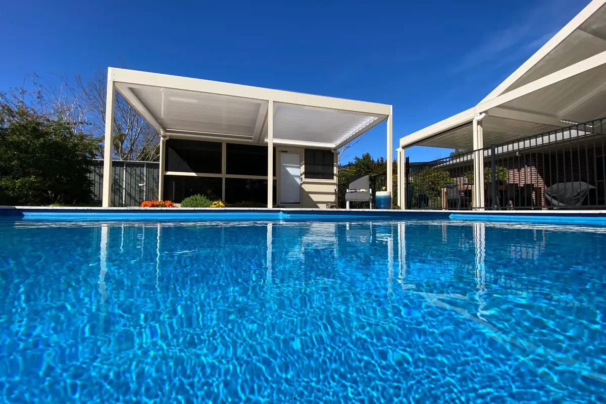 Freestanding louvred roof across the pool from a gabled patio, both in a cream powder-coat finish