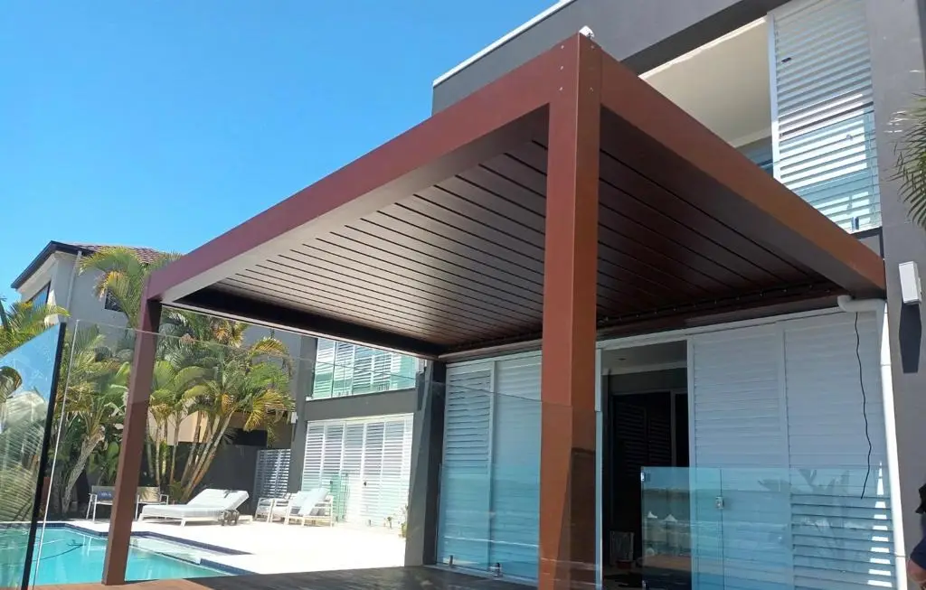 Louvred roof in a woodgrain powder-coat finish over a deck, blades closed, beside a glass-fenced pool
