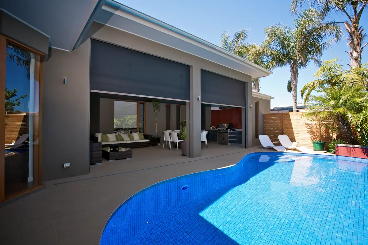Blinds part-lowered across a wide alfresco opening, seen from the far side of the pool