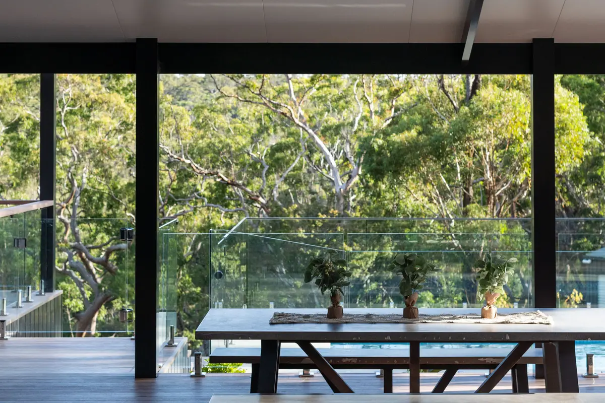 Deck boards running the length of the alfresco to a concrete dining table, bushland beyond the glass balustrade