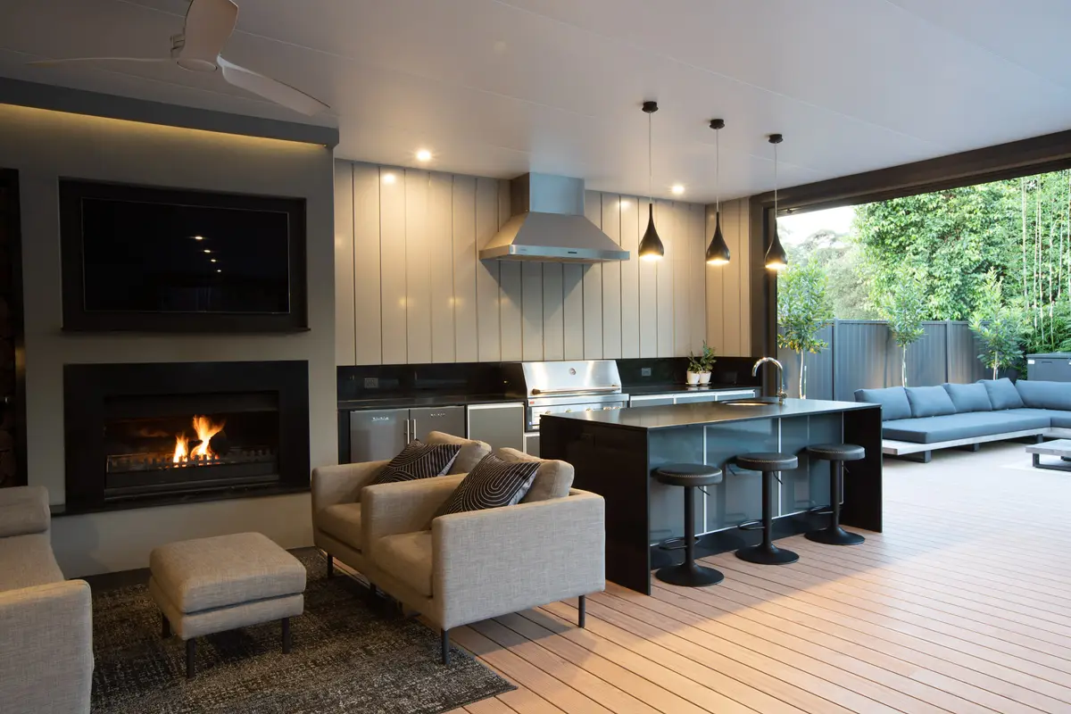 Clad wall behind the outdoor kitchen and range hood, with the fireplace lit at dusk