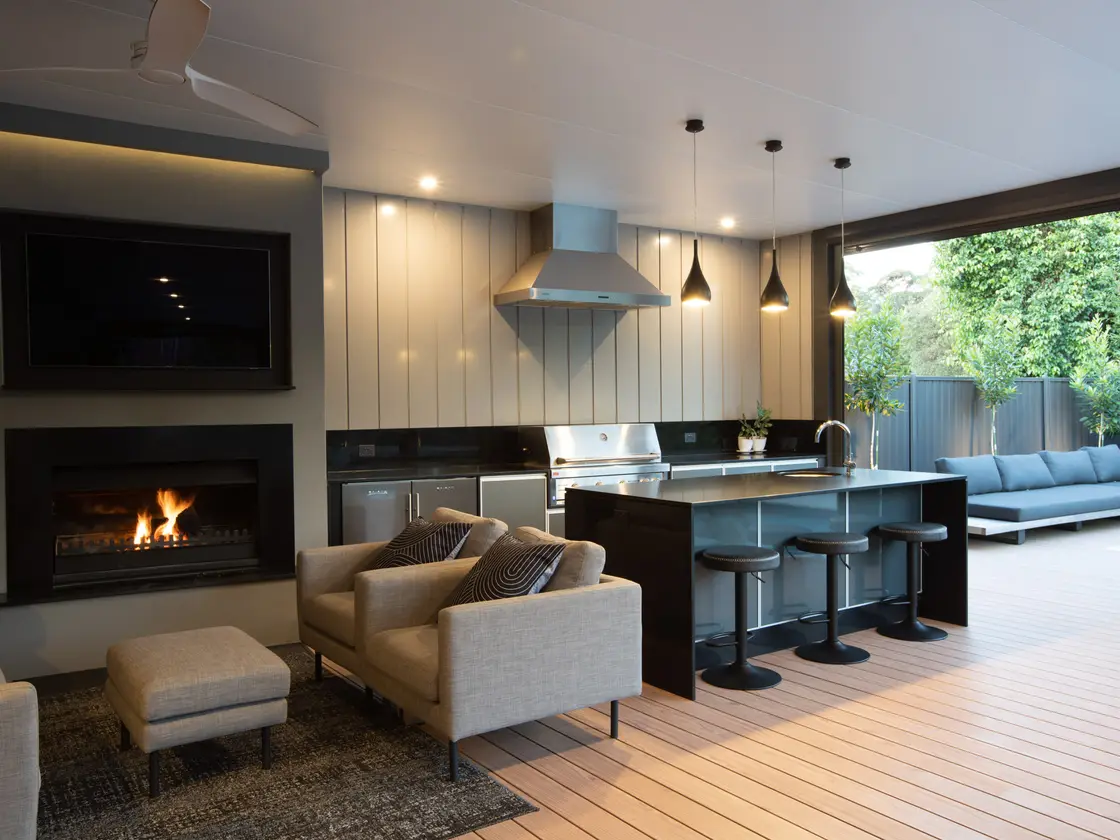 Screen recessed into a pale clad wall above a lit fireplace, outdoor kitchen and range hood alongside at dusk