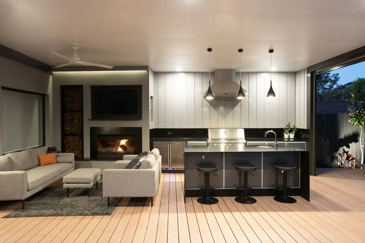 Outdoor kitchen backed by a white clad wall with the range hood run up through it, fireplace and TV alongside
