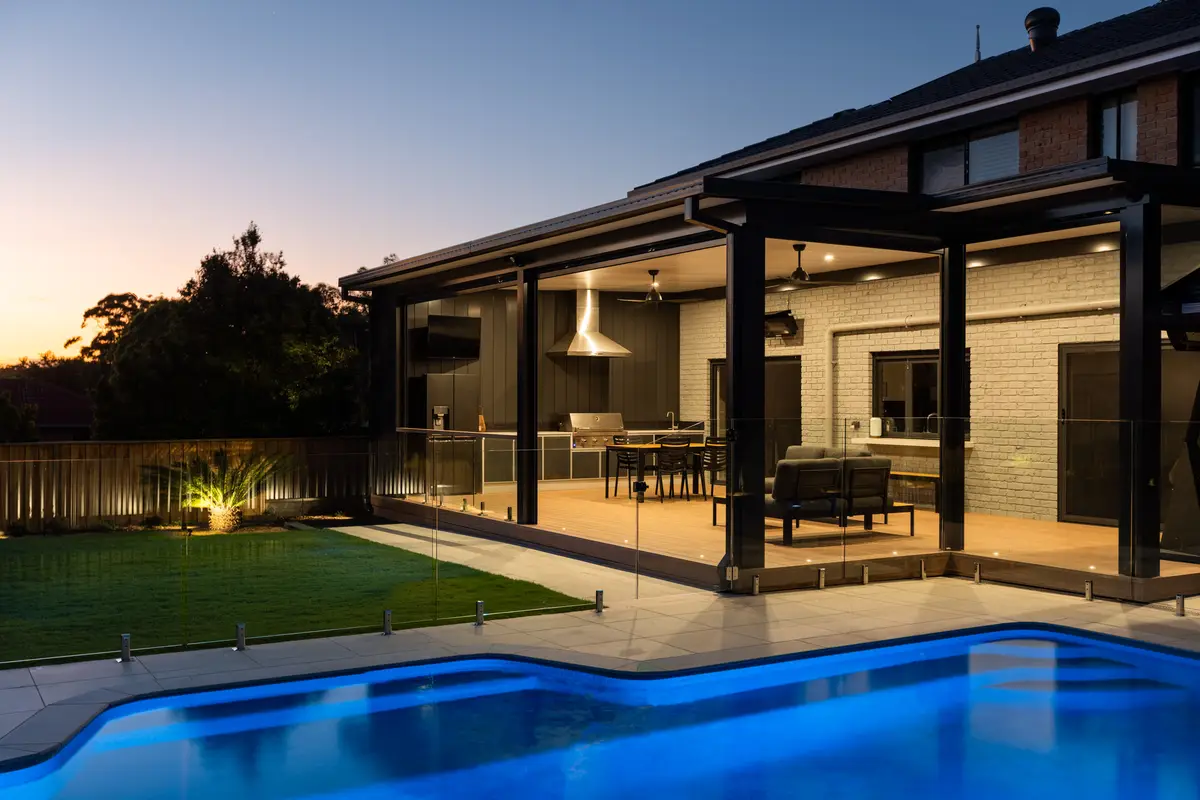 Clad wall behind the lit outdoor kitchen, seen across the pool at last light